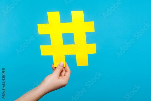 Closeup of female hand holding big yellow paper hashtag sign, hash symbol of internet popularity, famous media trends, blogging and viral content. indoor studio shot isolated on blue background