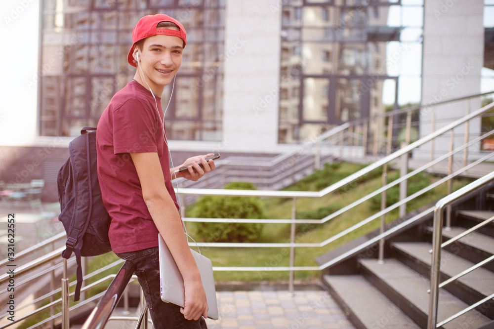 Smiling teenager holds cell phone and laptop, in the city.  Young man stands with a smartphone and computer, in the street.  Lifestyle concept of a happy teenage boy, outdoors.