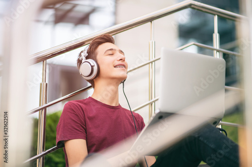 Smiling teenage boy with a laptop on the street. Handsome young man works on a notebook, outdoors. Dreaming guy holds a laptop on the knees and dreaming. Teenager in headphones with laptop