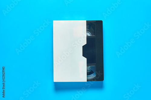 One black old vhs video cassette in white cardboard packaging on blue desk. Concept of 90s. Top view. Close-up