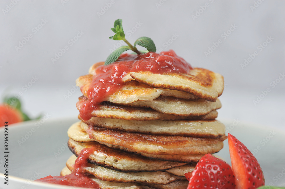 Pancakes with strawberry syrup. Fritters are decorated with fresh strawberries and mint leaves. Light background. Close-up. Pancakes with strawberry syrup. Fritters are decorated with fresh strawberries and mint leaves. Light background. Close-up.