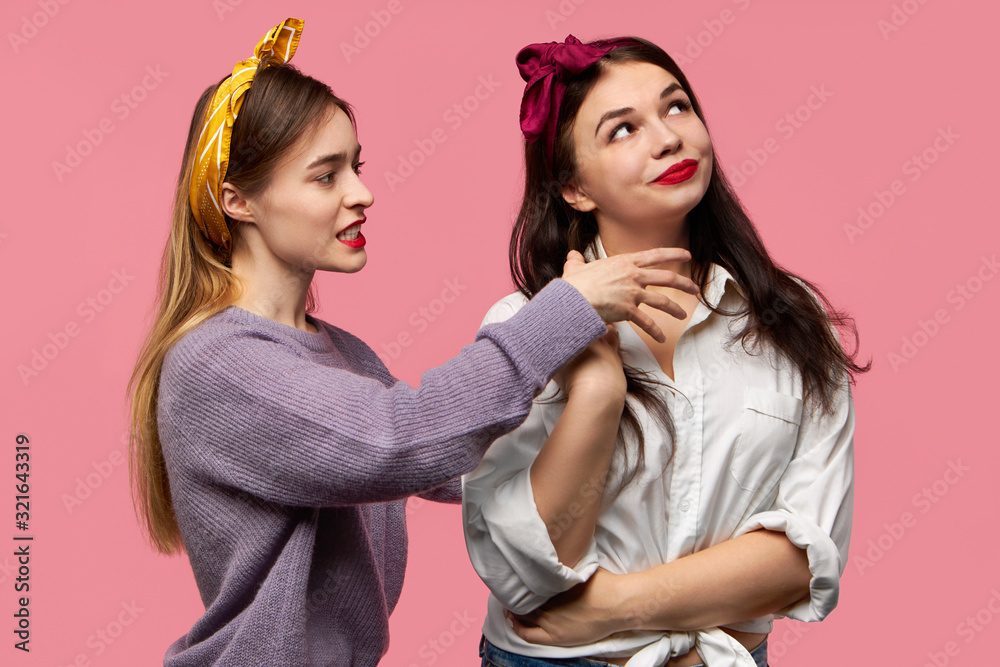 Emotional infuriated young female grimacing and screaming expressing negative reaction being angry holding hand on neck of her girlfriend with malevolent gloating smile as if trying to strangle her