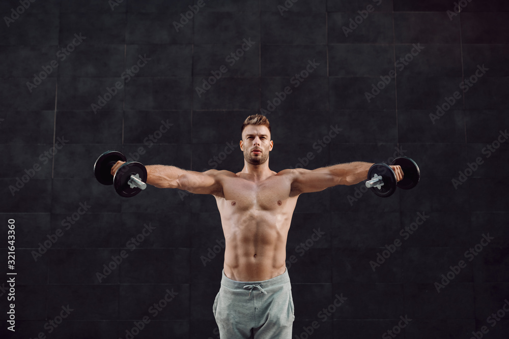 Attractive caucasian muscular bearded blonde shirtless man in tracksuit and standing in front of gray wall and holding dumbbells.
