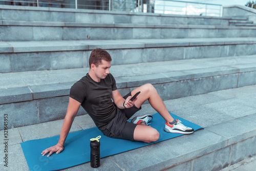 male athlete, in summer city, resting after workout, holding a smartphone, reading and writing message on smartphone, resting on a fitness mat. Bottle shaker with protein and water.