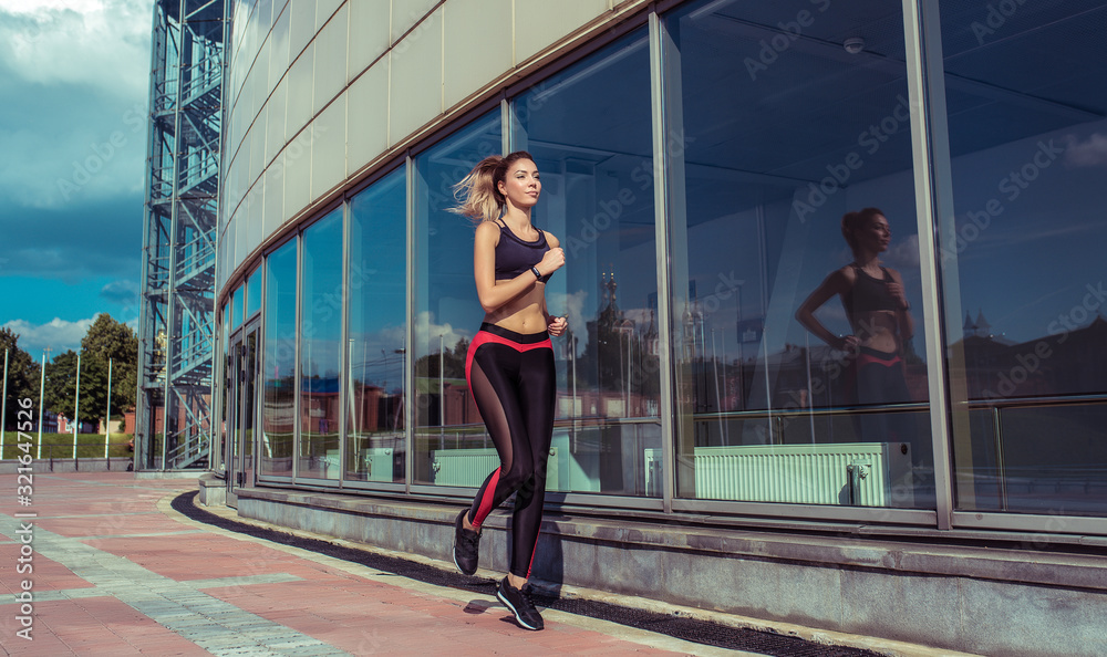 Beautiful girl, summer in city, fitness training on street, jogging in morning, sportswear, leggings top. Tanned figure. Movement in jump. Background glass windows of building. Free space copy text.