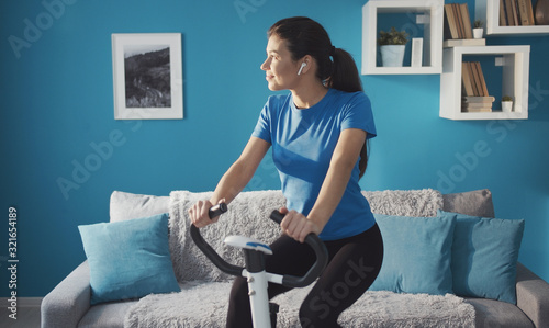 Sporty happy girl training on exercise bike at home listening music looking aside, healthy life
