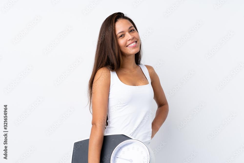 Young Brazilian girl holding a scale over isolated background with arms at hip and holding weighing machine Young Brazilian girl holding a scale over isolated background with arms at hip and holding weighing machine
