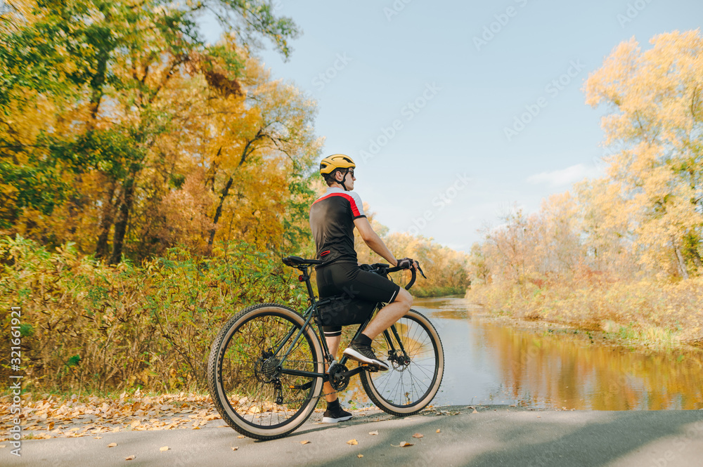 Obraz premium Photo of a cyclist standing on the shore of an autumn lake and resting, wearing a cycling jersey and helmet. Young man with a bike walking in a beautiful park. Cycling is a hobby.