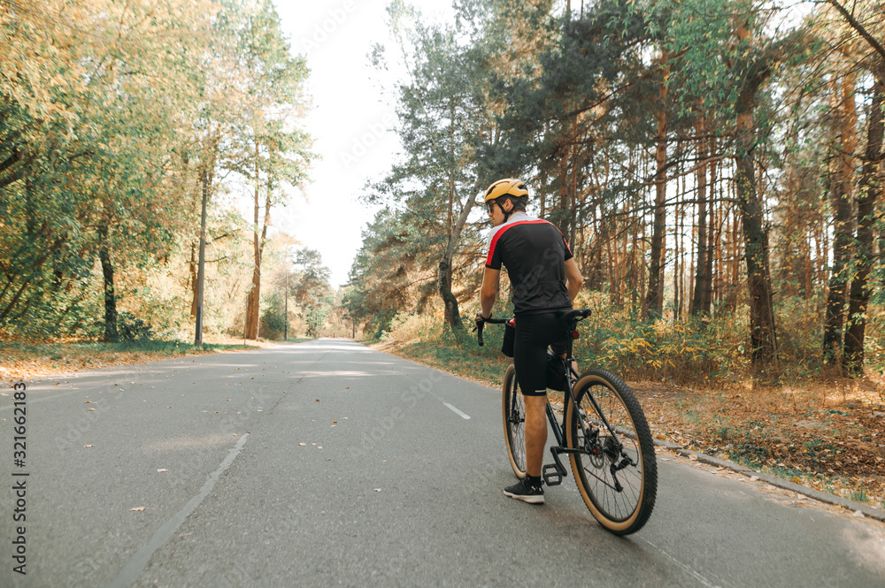 Cyclist in sportswear and helmet stands on the road in the forest and looks to the side, rear view. Cyclist on a walk in the woods with a bicycle in autumn day. Cycling workout in the park.