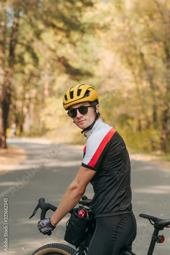 Portrait of handsome man in sunglasses and helmet standing with bicycle on road and looking into camera with serious face against background of autumn landscape of trees and road. Cyclist is walking