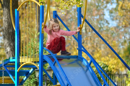 Little fair-haired girl has fun on a slide at the playground on a warm autumn day. The concept of a happy carefree childhood.