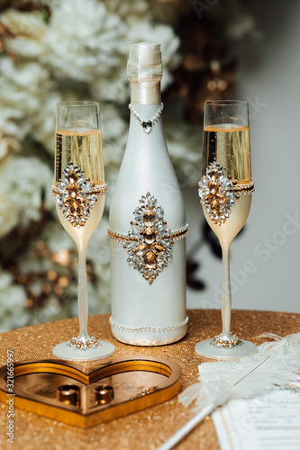 Two gold wedding rings lie in a heart-shaped box, a champagne bottle with glasses stands beside it. Jewelry, engagement ceremony, traditions concept.