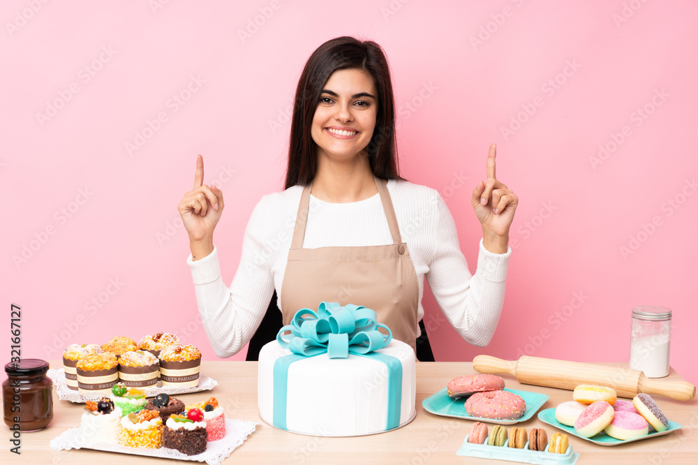 Pastry chef with a big cake in a table over isolated pink background pointing up a great idea