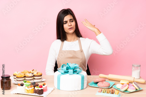 Pastry chef with a big cake in a table over isolated pink background with tired and sick expression