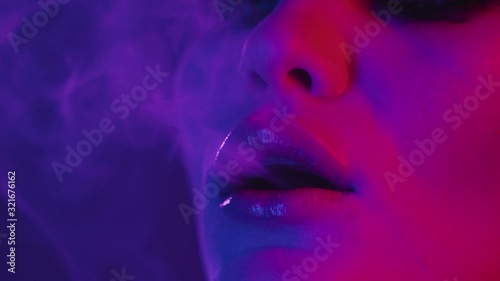 Colorful portrait of beauty sexy model girl in neon, blowing smoke of electronic cigarette or vape. Focused close-up of young woman face with dark make-up in UV pink and blue lights. Slow motion
