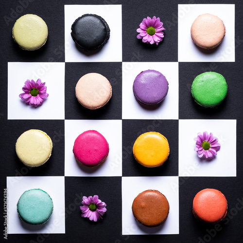 Creative layout of colorful macaroons and flowers on chessboard background. Variety of french almond macaron cookies.Dessert, sugar, bakery creative, playful concept.Alternatvie chess game.Food design