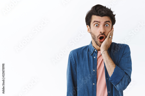 Shocked, speechless young handsome man with beard, gasping drop jaw and stare camera impressed, heard shocking rumor standing impressed white background, touch cheek after his crush kissed him