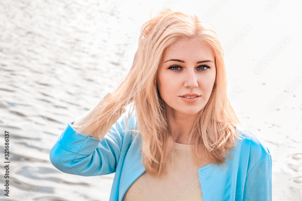 Portrait of an attractive young blonde woman in blue jacket on a blurred background, the woman looks at the camera, touching hair