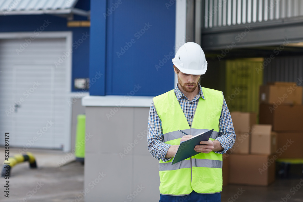 Young man in protective workwear standing and making notes in document while delivering load he working outdoors Young man in protective workwear standing and making notes in document while delivering load he working outdoors