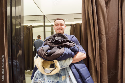 Young man with a armful of clothes in the fitting room during the sale and discounts