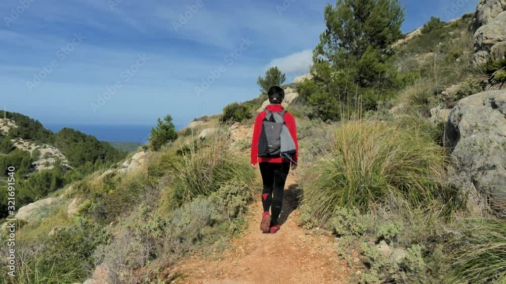 Woman backpacker  hiking within mountain trek on Mallorka islan. Sunny spring  day with comfortable temperature. Rocky sandy footpath, pine trees, bushes and palms around.