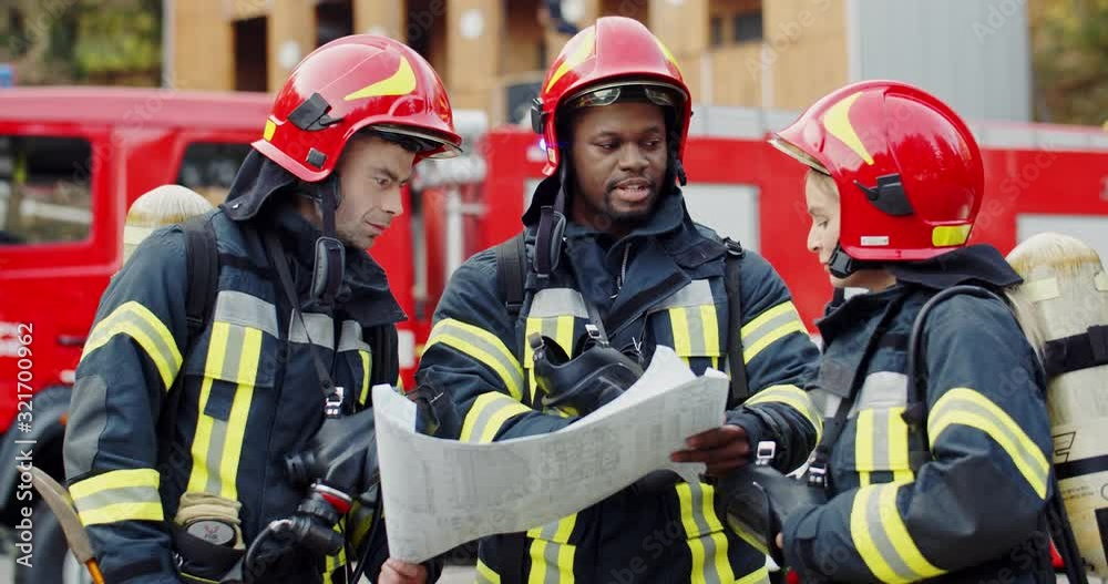 Caucasian pretty woman and wo multiethnic men fire fighters co-workers standing outdoors at the fire truck with a big plan or map in hands and talking while deciding how to fight with fire.