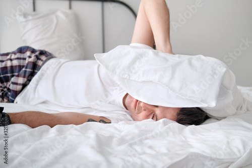 Young man in the white shirt lying on a bed with white linen and covers his head with a pollow