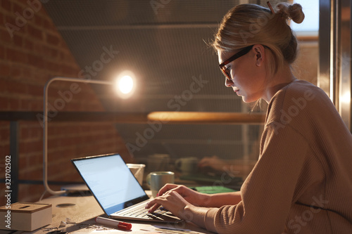 Young blonde woman in eyeglasses and in casual clothing sitting at desk and working on laptop till late at office