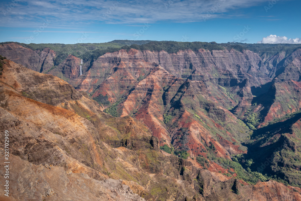 Waimea Canyon, Kauai, Hawaii, USA. - January 16, 2020: Wide landscape with waterfall in distance shows red rocks partly green in crevasses, green cover on top, under blue sky. Waimea Canyon, Kauai, Hawaii, USA. - January 16, 2020: Wide landscape with waterfall in distance shows red rocks partly green in crevasses, green cover on top, under blue sky.