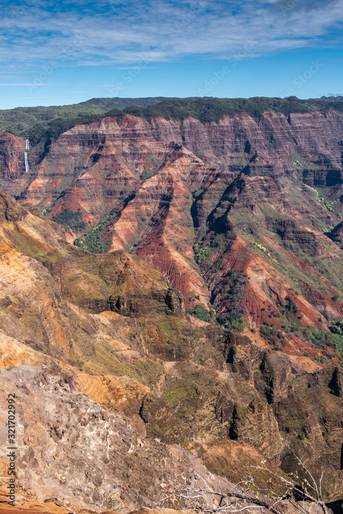 Waimea Canyon, Kauai, Hawaii, USA. - January 16, 2020: Portrait of double white waterfall in distance among red rocky side and with green tree cover on top under blue sky. Waimea Canyon, Kauai, Hawaii, USA. - January 16, 2020: Portrait of double white waterfall in distance among red rocky side and with green tree cover on top under blue sky.