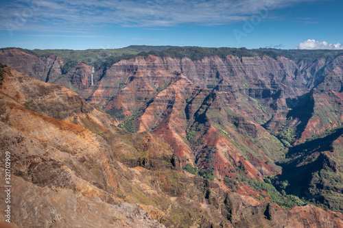 Waimea Canyon, Kauai, Hawaii, USA. - January 16, 2020: Wide landscape with waterfall in distance shows red rocks partly green in crevasses, green cover on top, under blue sky.