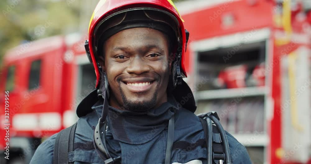 Close up of the cheerful African American young fireman in the helmet smiling to the camera after fire at the red truck. Portrait. Close up of the cheerful African American young fireman in the helmet smiling to the camera after fire at the red truck. Portrait.