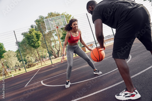 Outdoors Activity. African couple guy dribbling back view close-up while girl blocking smiling joyful on basketball court