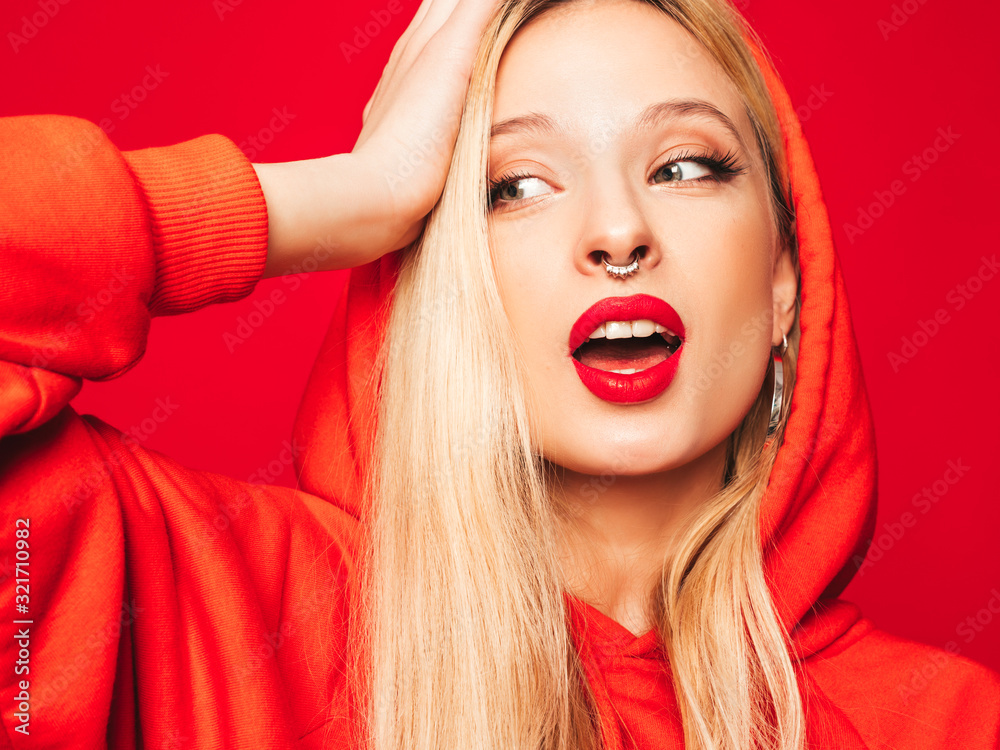 Portrait of young beautiful hipster bad girl in trendy red summer hoodie and earring in her nose.Sexy carefree smiling blond woman posing in studio.Positive model having fun Portrait of young beautiful hipster bad girl in trendy red summer hoodie and earring in her nose.Sexy carefree smiling blond woman posing in studio.Positive model having fun