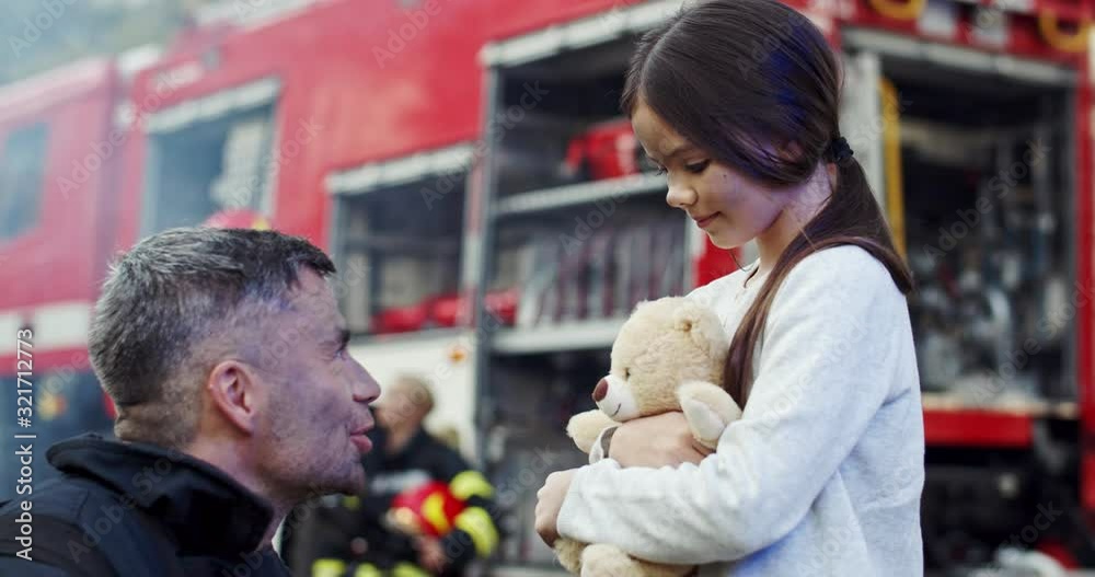 Good Caucasian fire fighter calming down little scared girl with a teddy bear which being saved by him from a fire. Good Caucasian fire fighter calming down little scared girl with a teddy bear which being saved by him from a fire.