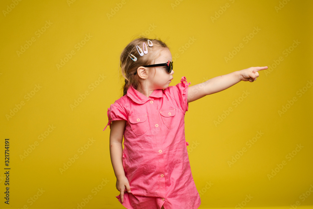 Stock photo of cute little girl with hair accessories wearing sunglasses and pink dress showing the direction with her index finger. Isolate on yellow background. Stock photo of cute little girl with hair accessories wearing sunglasses and pink dress showing the direction with her index finger. Isolate on yellow background.