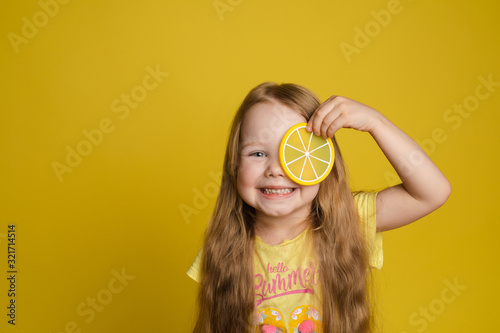 Tableau sur toile Portrait of smiling cute little girl holding slice of lemon near eye looking at camera medium close-up