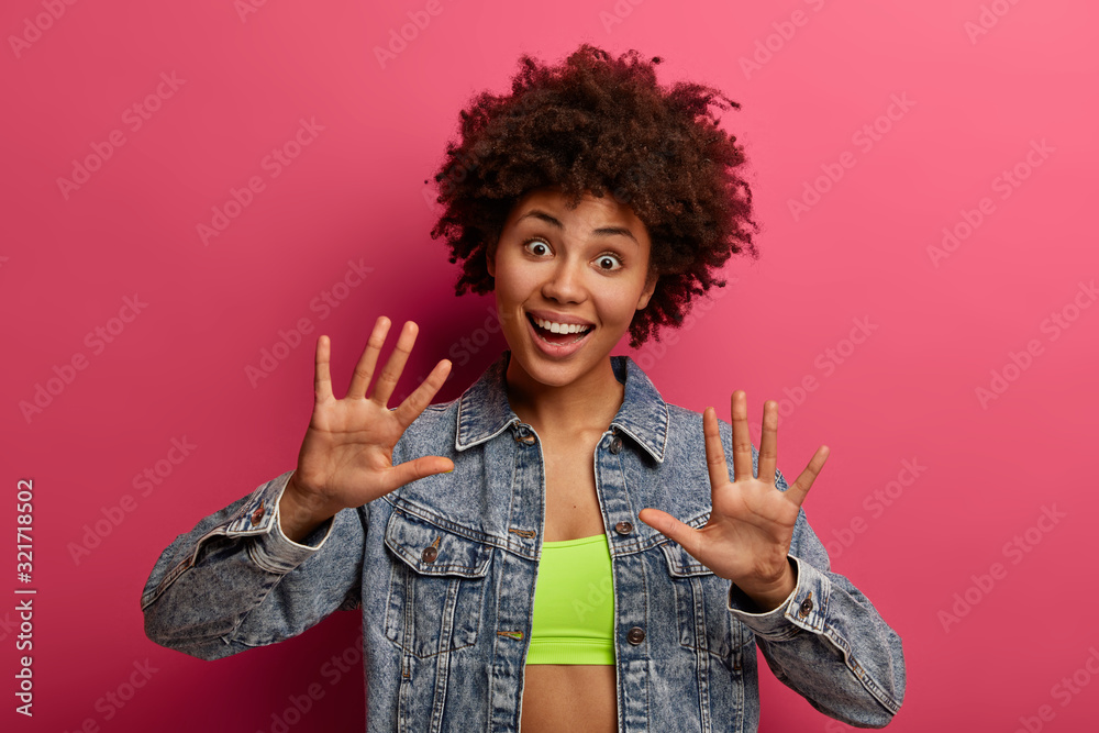Joyful happy woman with dark skin, curly hair, broad smile, extends palms at camera, wears fashionable denim jacket and top, has surprised expression, looks with funny playful expression, poses indoor Joyful happy woman with dark skin, curly hair, broad smile, extends palms at camera, wears fashionable denim jacket and top, has surprised expression, looks with funny playful expression, poses indoor