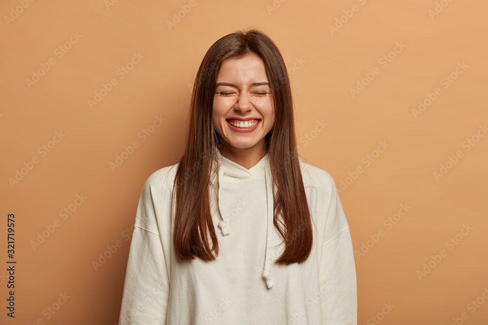 Overemotive brunette woman has beaming smile, feels amused, hears hilarious story, closes eyes and shows white ideal teeth, dressed in casual hoodie, has happy chuckle, isolated on brown wall Overemotive brunette woman has beaming smile, feels amused, hears hilarious story, closes eyes and shows white ideal teeth, dressed in casual hoodie, has happy chuckle, isolated on brown wall