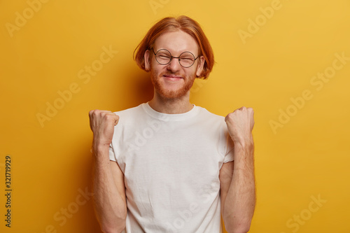 Isolated shot of happy triumphing man raises clenched fists, closes eyes, celebrates success and victory, has ginger hair, beard, wears spectacles and white t shirt, isolated over yellow background