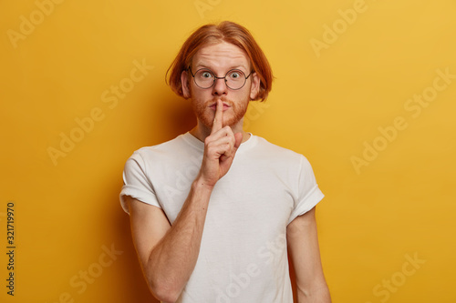 Portrait of mysterious teenager has bob hairstyle, ginger beard, presses index finger over lips, makes hush gesture, gossips about something very secret, asks nor spread rumors, dressed casually