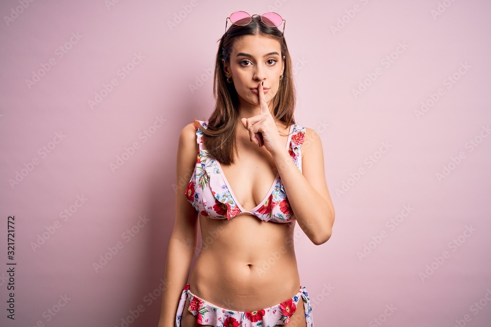 Fototapeta premium Young beautiful woman on vacation wearing bikini and sunglasses over pink background asking to be quiet with finger on lips. Silence and secret concept.