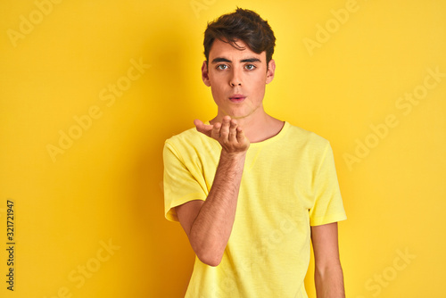 Teenager boy wearing yellow t-shirt over isolated background looking at the camera blowing a kiss with hand on air being lovely and sexy. Love expression.