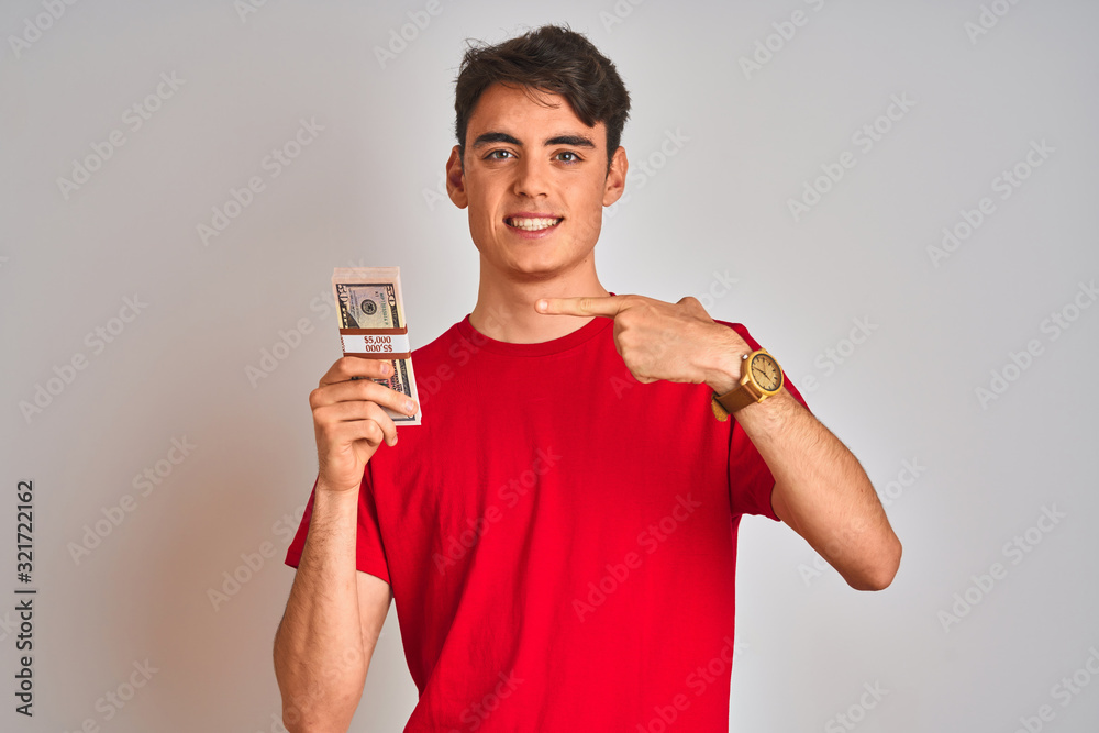 Teenager boy holding a bunch of dollars bank notes over isolated background very happy pointing with hand and finger Teenager boy holding a bunch of dollars bank notes over isolated background very happy pointing with hand and finger