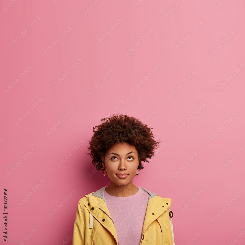 Thoughtful black woman with curly hair looks attentively above, notices something interesting, dressed casually, has mysterious face expression, isolated over pink pastel background. Hmm let me think Thoughtful black woman with curly hair looks attentively above, notices something interesting, dressed casually, has mysterious face expression, isolated over pink pastel background. Hmm let me think