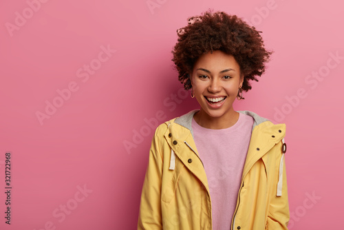 Outgoing optimistic millennial girl has fun and smiles toothily, being in high spirit, dressed in casual wear, chuckles from funny joke, stands over rosy wall with copy space for your advertisement