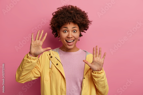Isolated shot of joyous curly woman keeps palms raised, enjoys free time, plays with someone, smiles broadly, looks at camera with happiness, wears yellow jacket, poses against rosy background