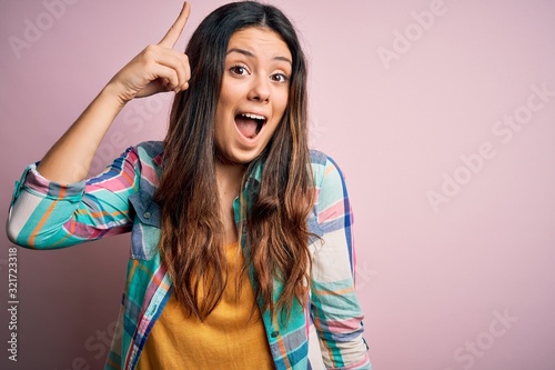 Young beautiful brunette woman wearing casual colorful shirt standing over pink background pointing finger up with successful idea. Exited and happy. Number one.