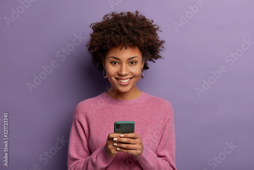 Positive dark skinned woman social media manager makes new content on smartphone, texts and smiles broadly, dressed in casual wear, models against purple background, watches video in internet