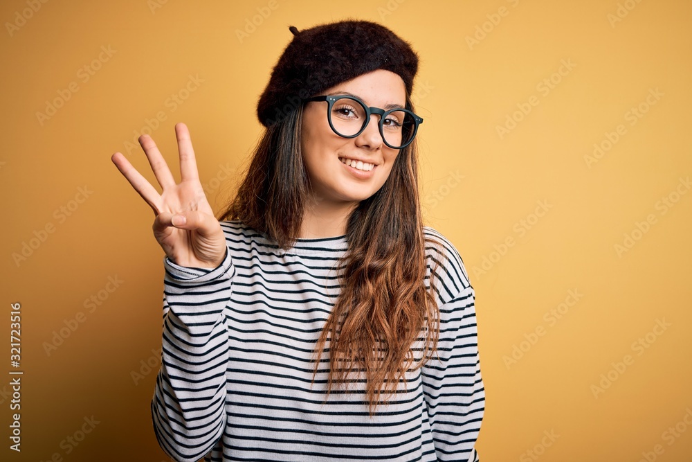 Young beautiful brunette woman wearing french beret and glasses over yellow background showing and pointing up with fingers number three while smiling confident and happy. Young beautiful brunette woman wearing french beret and glasses over yellow background showing and pointing up with fingers number three while smiling confident and happy.